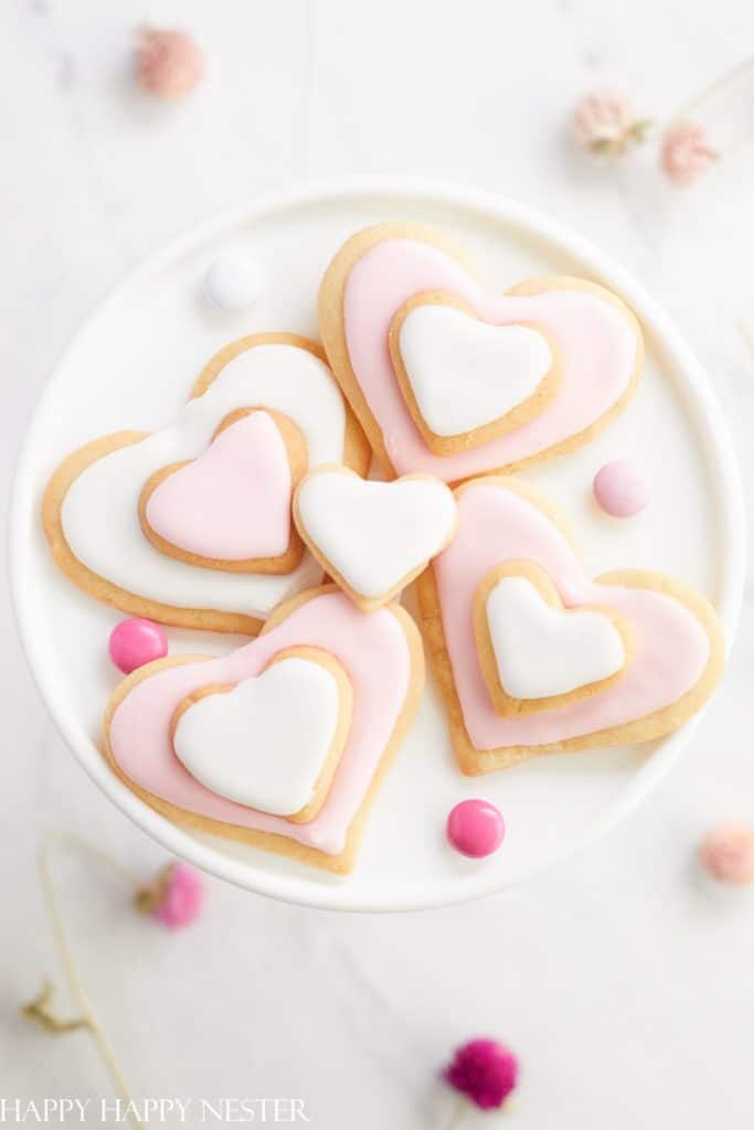 Heart-shaped cookies with white and pink icing are arranged on a cake stand, surrounded by pink and white candies. These delightful pink desserts are perfectly accented by dried flowers scattered around the pristine white surface.