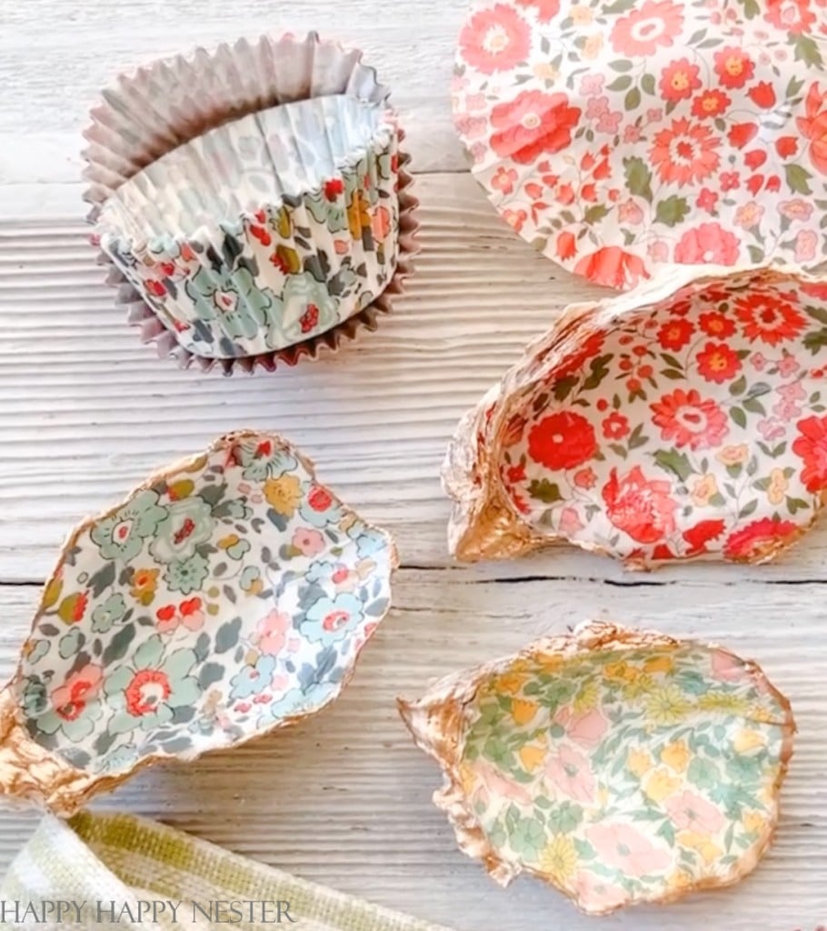 Decorative pink floral-patterned paper cupcake liners are displayed alongside oyster shells lined with similar floral paper, set on a light wooden surface. A green striped cloth is partially visible in the foreground, adding a vibrant touch to these delightful crafts.