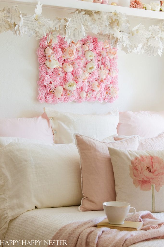 A cozy white and pink bed setup with fluffy pillows and a throw blanket. Above the bed, a decorative square panel made of pink and white roses showcases intricate pink crafts, while a floral garland hangs on a white shelf. A white mug sits on the bed.