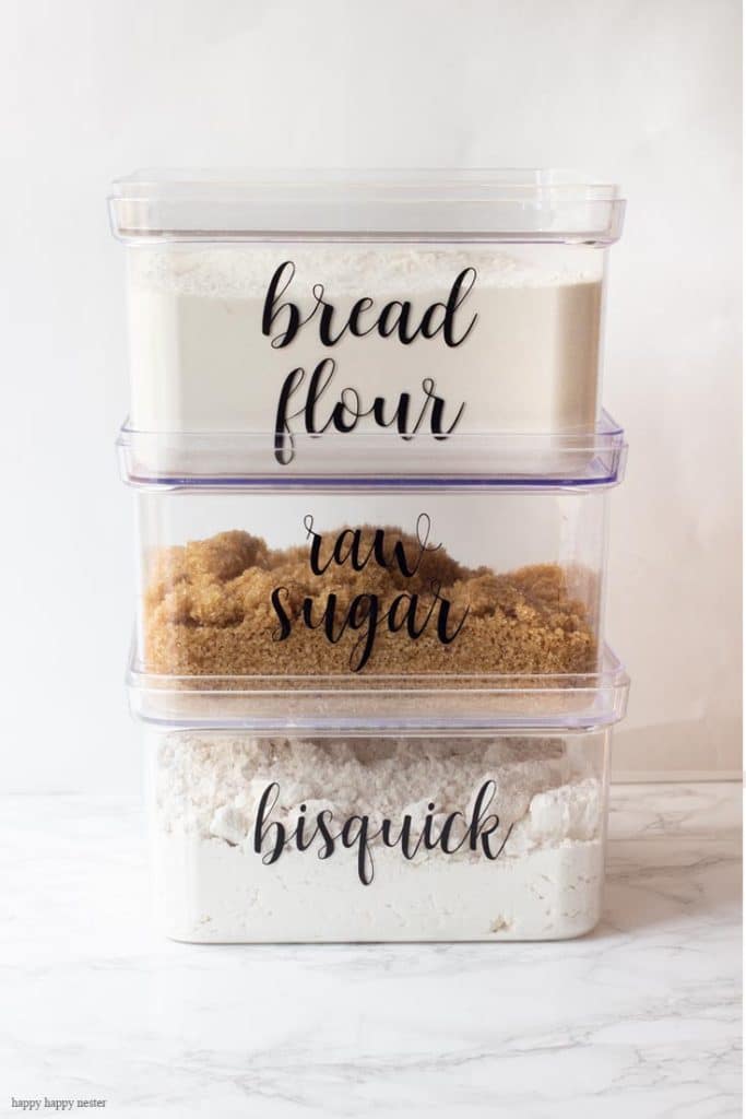 Three clear containers are stacked vertically, showcasing professional organizing tips. The top holds bread flour, the middle cradles raw sugar, and the bottom contains Bisquick. Each container is labeled in black cursive lettering and rests gracefully on a white marble surface.