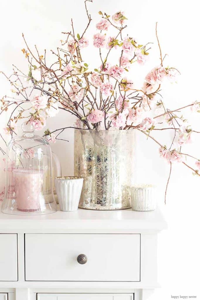 A white dresser adorned with a vase of blooming pink cherry blossoms exudes charm. This artistic display, akin to pink crafts, is completed by two white candles in textured holders and a clear pitcher alongside, creating a delicate and serene scene against the pure backdrop.