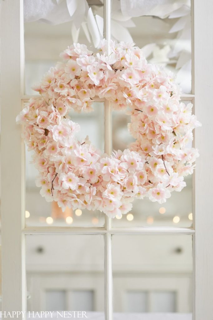 A charming wreath crafted from soft pink cherry blossoms hangs on a glass door. The background is blurred, showcasing a softly lit interior with twinkling lights and adding a touch of whimsy to the pink crafts display.