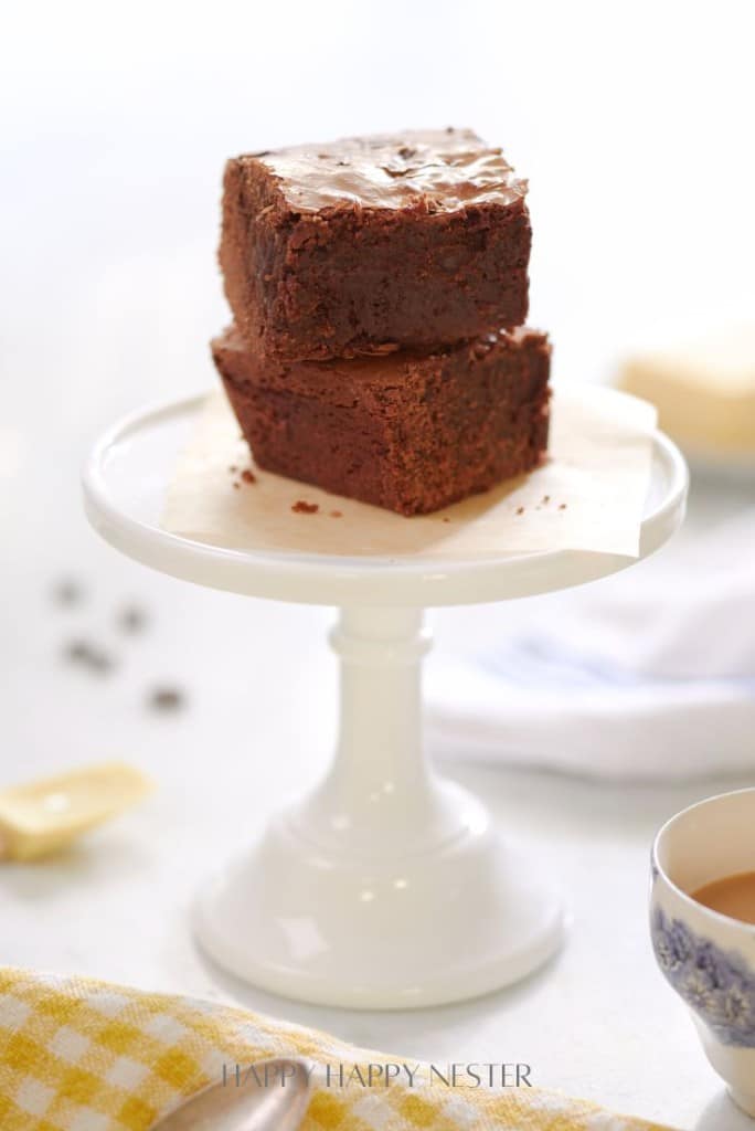 Two indulgent double chocolate brownies stacked on a white pedestal stand. A piece of parchment paper is underneath the brownies. A blurred yellow-checkered cloth and floral-patterned cup are partially visible in the background, hinting at the cozy scene of a beloved brownie recipe.