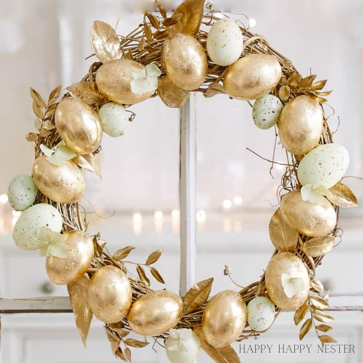 A decorative seasonal wreath adorned with gold and pastel-colored eggs, interspersed with gold leaves. The wreath is hung on a light background, with a soft, blurred effect in the distance.