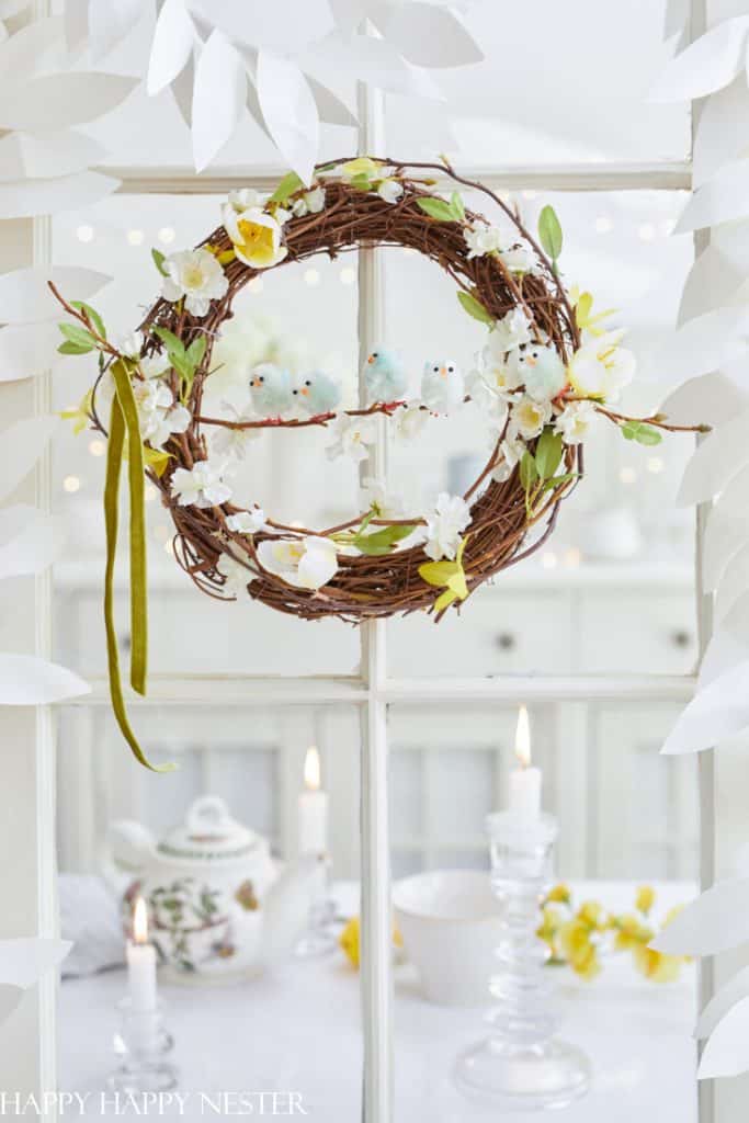 A decorative spring wreath hangs on a window, adorned with white and yellow flowers and small bird ornaments. Below, a table showcases seasonal wreaths, a floral teapot, burning candles in glass holders, and a bowl. White paper leaves cascade around.