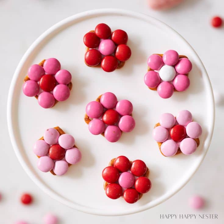 A white plate holds seven flower-shaped candy arrangements, reminiscent of pink desserts. Each consists of red, pink, and purple candies assembled in petal patterns on a circular base. The background is soft and blurred.