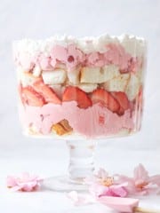 Indulge in this easy fruit trifle recipe featuring a layered dessert in a glass bowl, complete with whipped cream, pink mousse, sliced strawberries, and chunks of cake. The creation is elegantly surrounded by light pink flower petals.