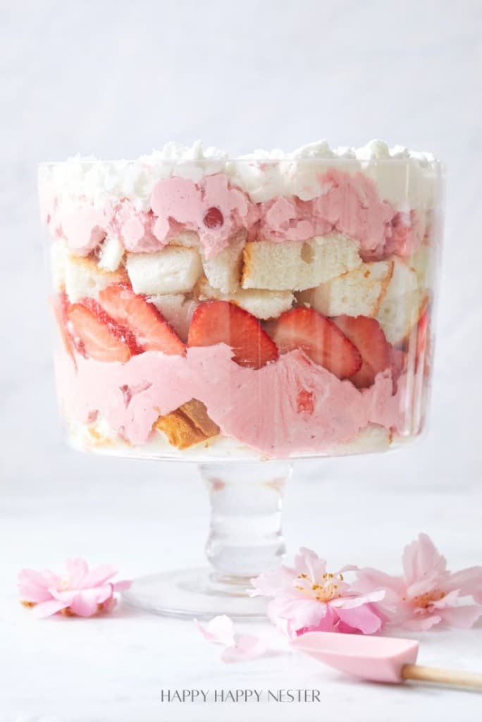 Indulge in this easy fruit trifle recipe featuring a layered dessert in a glass bowl, complete with whipped cream, pink mousse, sliced strawberries, and chunks of cake. The creation is elegantly surrounded by light pink flower petals.