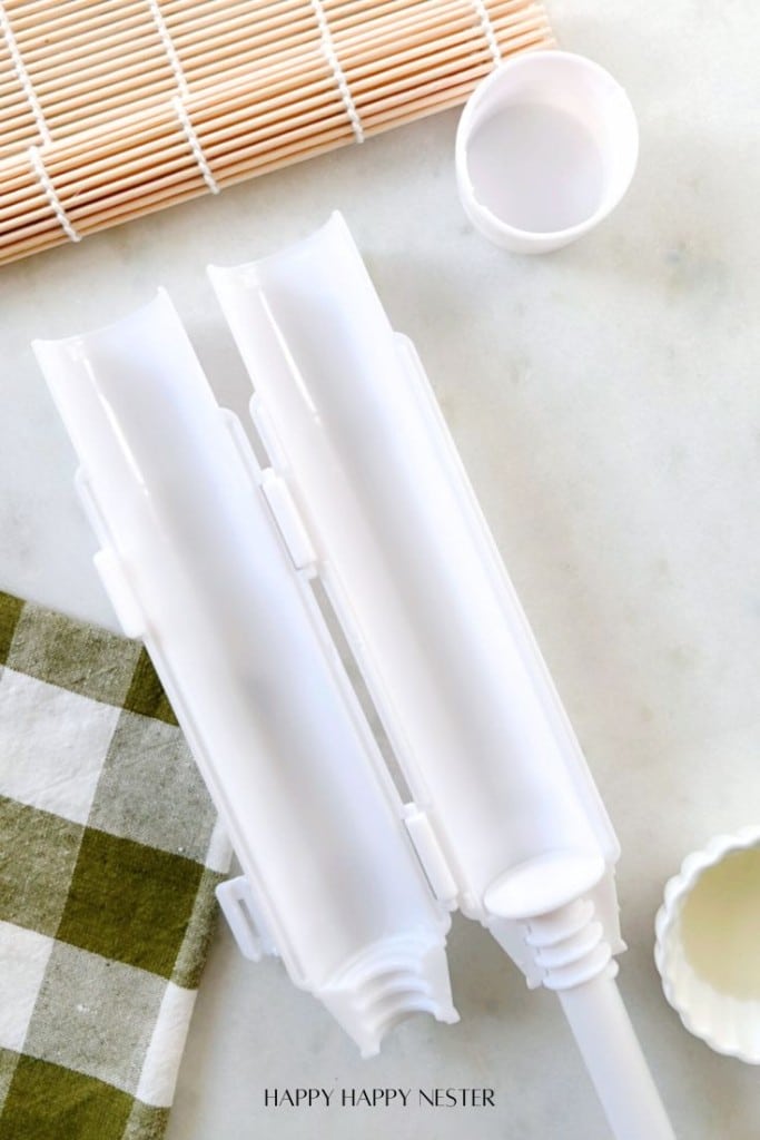 A sushi bazooka placed on a marble countertop next to a bamboo sushi mat, a small round container, and a green and white checkered cloth.