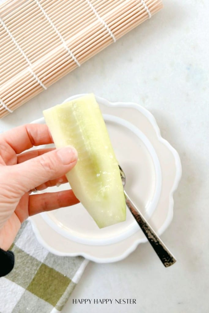 A hand holds a peeled cucumber slice over a decorative white plate and spoon. A bamboo mat and checkered cloth are in the background.