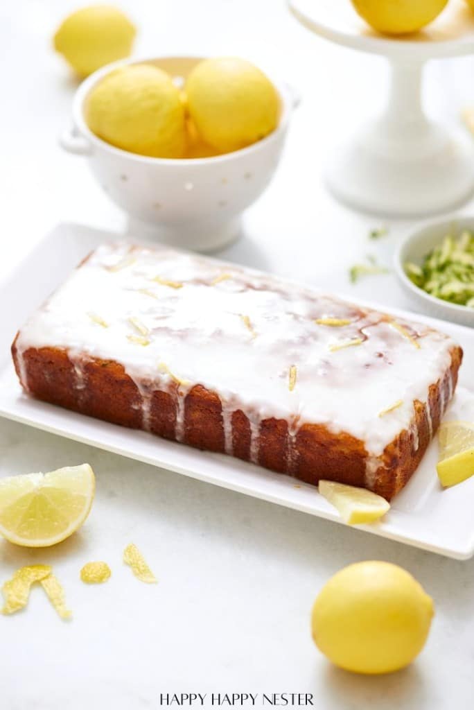 A rectangular healthy lemon loaf with white glaze sits on a white platter. Fresh lemons and slices surround it, with grated zest in a small bowl nearby. A colander filled with lemons is in the background, creating a bright, fresh scene perfect for any lemon loaf recipe enthusiast.