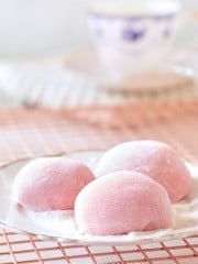 A plate with three pink mochi desserts covered in powdered sugar, placed on a wire cooling rack. A blurred teacup with blue patterns is in the background.