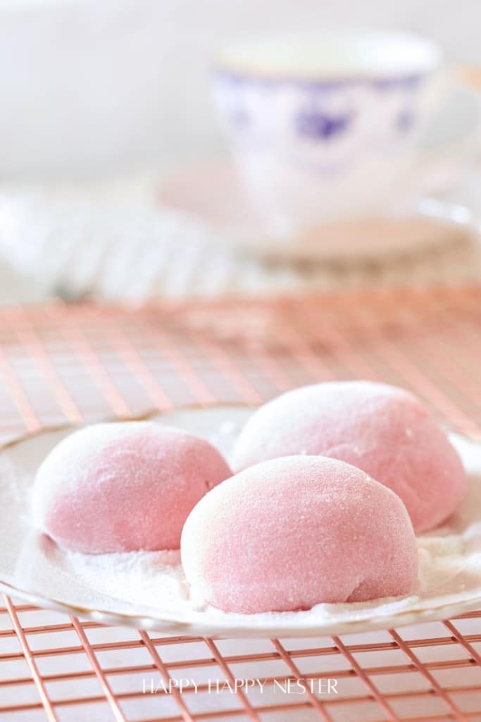 A plate with three pink mochi desserts covered in powdered sugar, placed on a wire cooling rack. A blurred teacup with blue patterns is in the background.