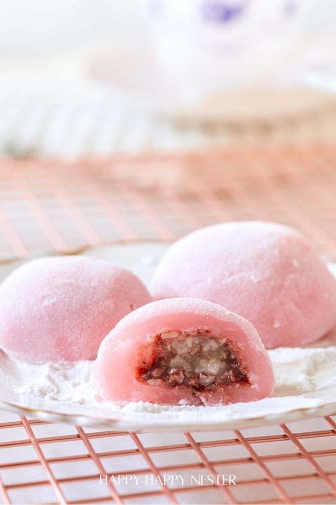 Close-up of three easy mochi desserts on a white plate. One is cut open, revealing a sweet red bean and nut filling. The plate sits on a pink wire rack against a soft focus background, showcasing these delightful treats.