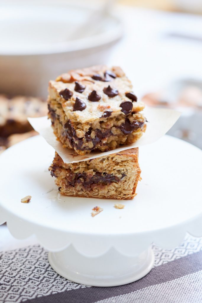 Two stacked square pieces of chocolate chip banana bread sit on a white plate, showcasing recipes made with chocolate chips. Each piece is layered with visible chocolate chips and a soft, moist texture, atop white paper. The background is softly blurred, revealing hints of kitchen elements.