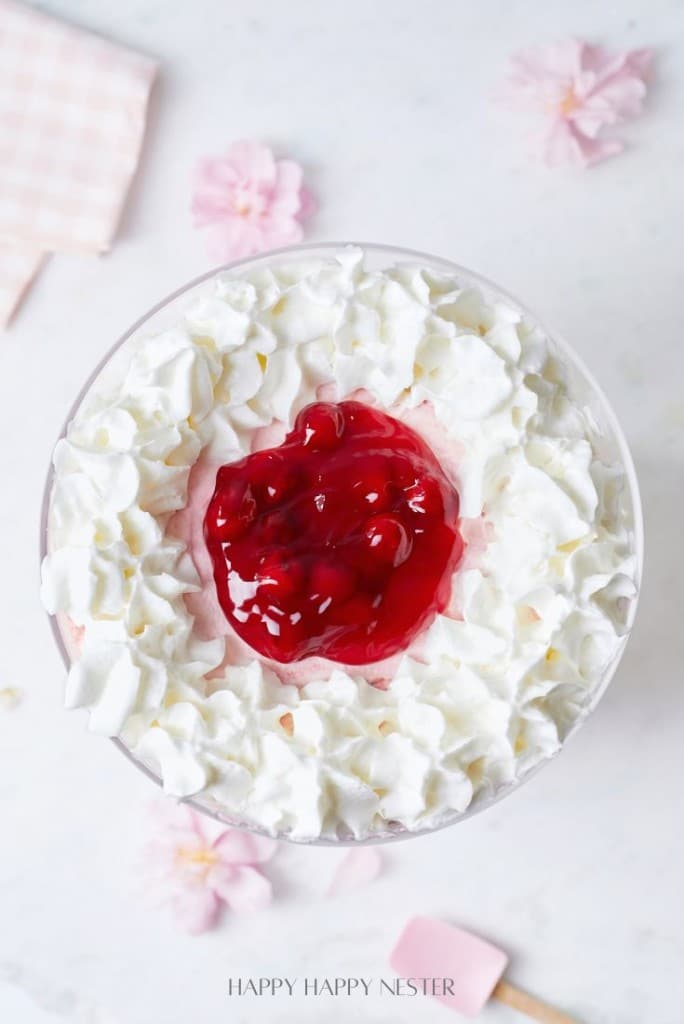 Top view of an easy fruit trifle dessert featuring a whipped cream border with a bright red cherry topping in the center. The tempting creation sits on a light surface, surrounded by pink flower petals and a checkered cloth, making it perfect for any gathering.