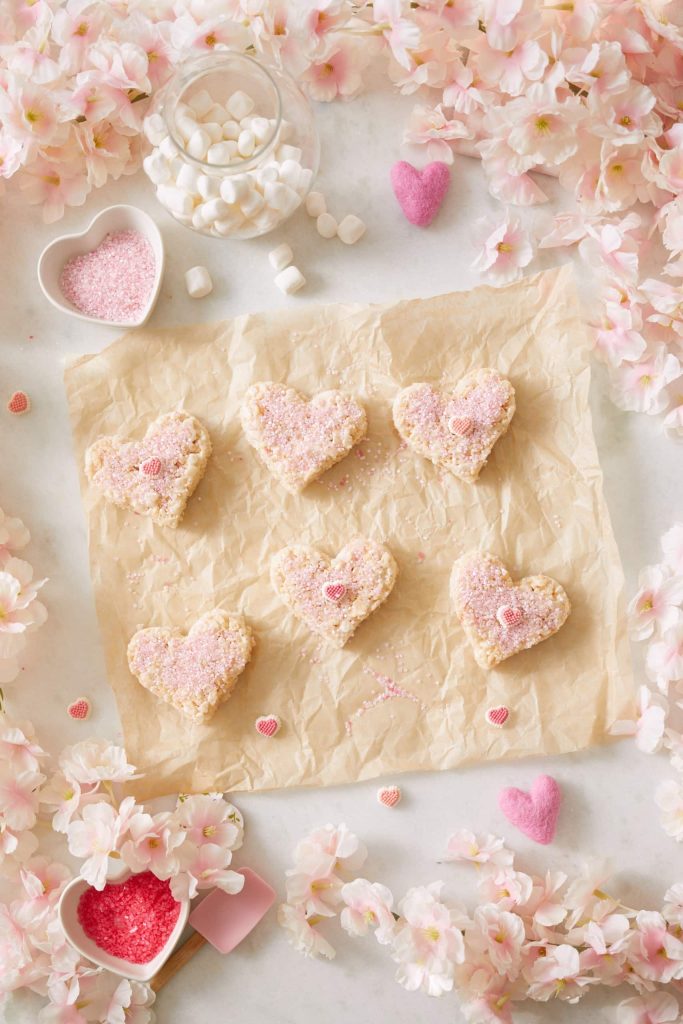 Heart-shaped pink desserts, adorned with sugar and tiny heart candies, are elegantly arranged on parchment paper. Encircling the scene are soft pink flowers, a dish of sugar, felt hearts, and a jar of marshmallows, crafting a romantic, pastel-themed setting.