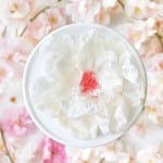 A white bowl holds a delicate white flower-shaped dessert with a pink center, surrounded by soft pink cherry blossom petals.