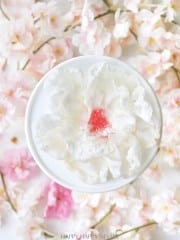 A white bowl holds a delicate white flower-shaped dessert with a pink center, surrounded by soft pink cherry blossom petals.