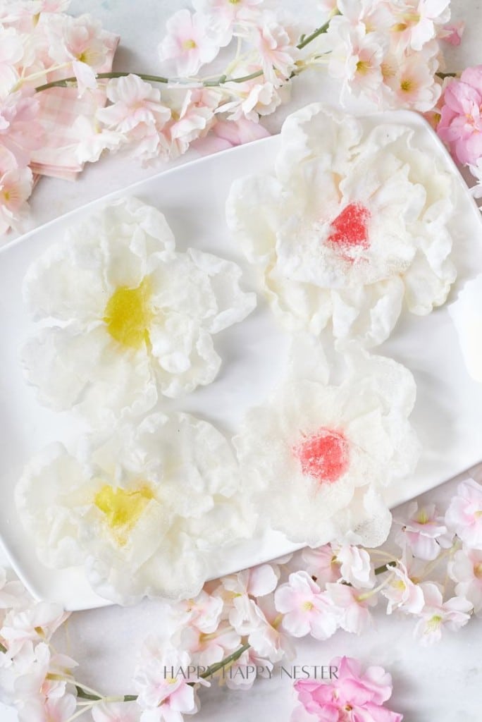 A white rectangular plate holds delicate fried rice paper flowers with translucent petals and hints of pink and yellow centers, surrounded by scattered pink and white blossoms on a marble surface.