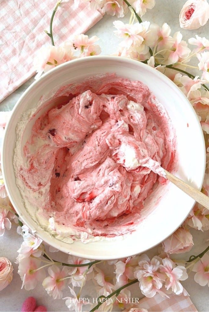 A bowl filled with pink whipped cream sits on a light surface, surrounded by delicate pink cherry blossoms. A wooden spoon is resting inside the bowl, and a pink checkered cloth is partially visible.