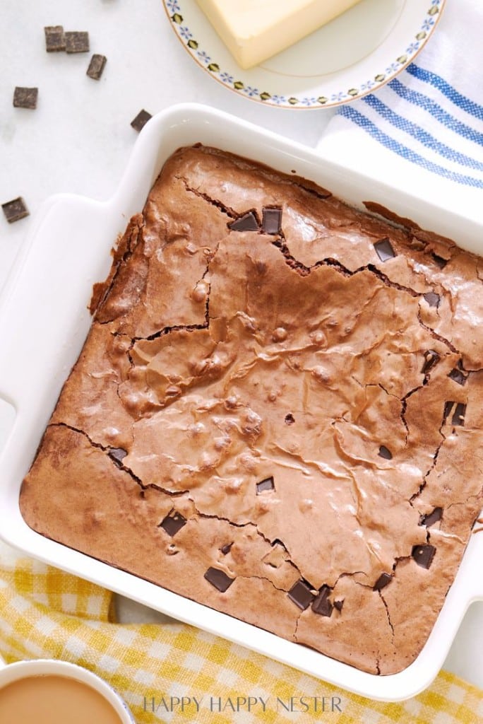 A freshly baked pan of brownies with a cracked, glossy top and visible chocolate chunks is placed on a white surface. It's surrounded by a blue-striped cloth, a butter dish, and a yellow-checkered towel.