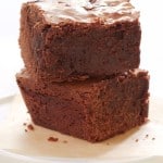 Two thick, fudgy brownies stacked on a white cake stand. The top brownie has a slightly cracked surface, showcasing a rich, chocolate texture. The plate sits on a napkin with faint blue stripes.