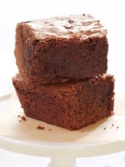 Two thick, fudgy brownies stacked on a white cake stand. The top brownie has a slightly cracked surface, showcasing a rich, chocolate texture. The plate sits on a napkin with faint blue stripes.