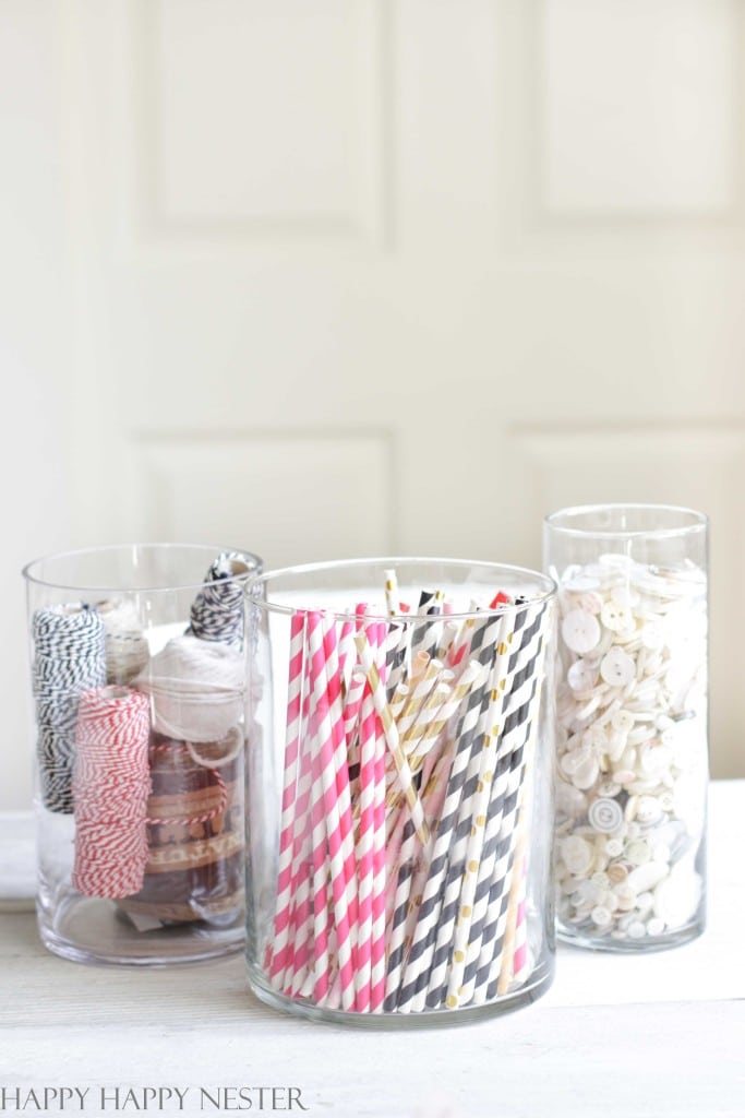 Three glass containers on a white surface showcase professional organizing tips: the left holds rolls of colorful twine, the center is filled with pink and black striped straws, and the right contains white buttons. A blurred background with a light-colored wall adds a touch of elegance.