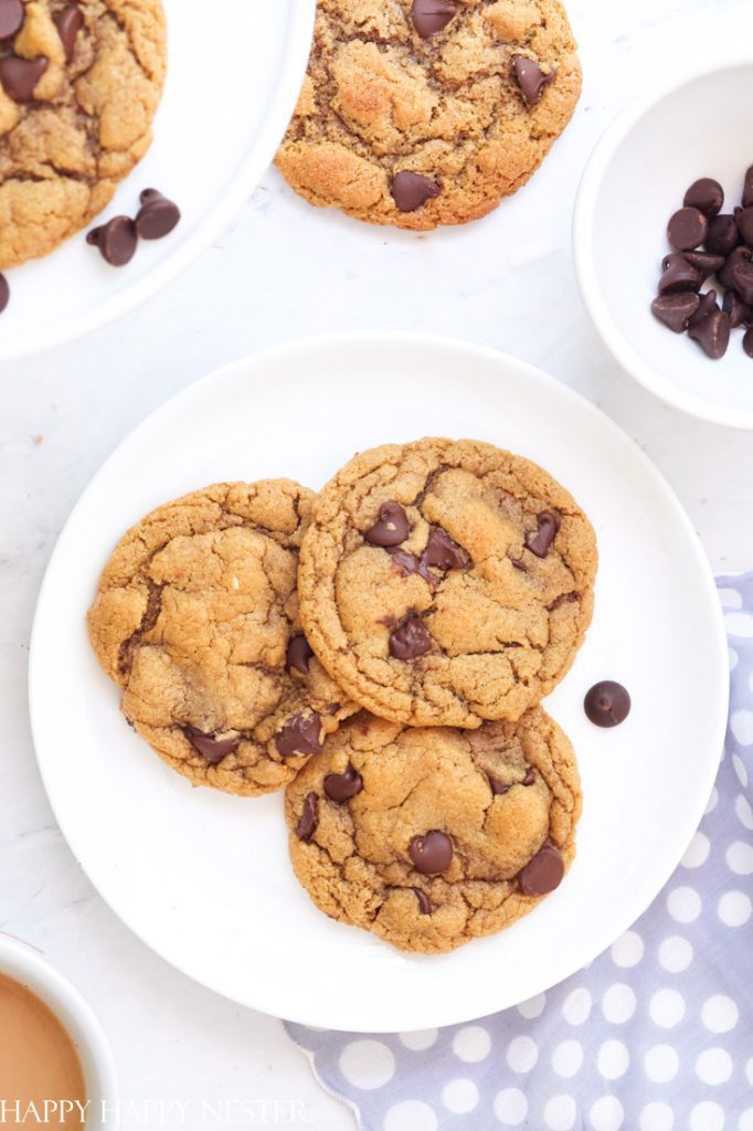 Three chocolate chip cookies sit on a white plate, surrounded by a few extra chips. Another plate of these irresistible treats is nearby. A polka-dotted napkin peeks out at the bottom, inviting thoughts of delightful recipes made with chocolate chips.