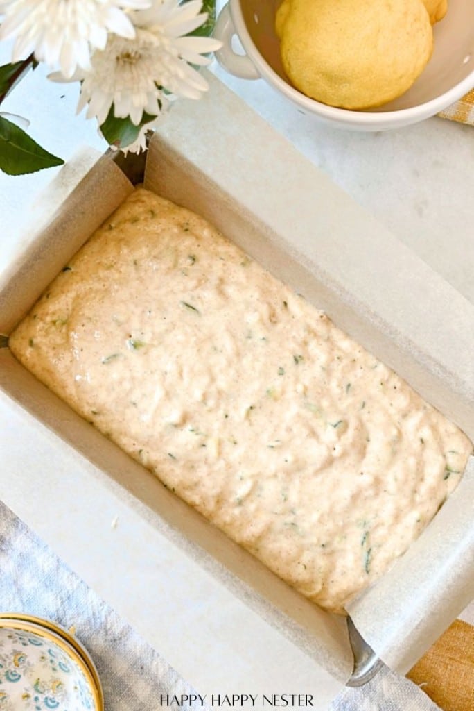 A loaf pan filled with uncooked zucchini bread batter sits on a countertop, ready to transform into a healthy lemon-infused loaf. Nearby, a white flower, small bowl of lemons, and patterned dish add charm to the setting.