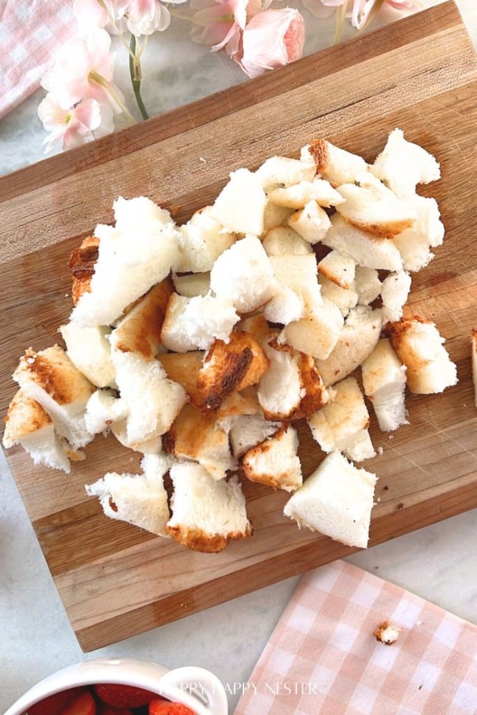 Cubes of torn white bread sit on a wooden cutting board, ready to accentuate an easy fruit trifle recipe. Pink flowers bloom in the background, while a checked pink cloth adds a touch of charm.