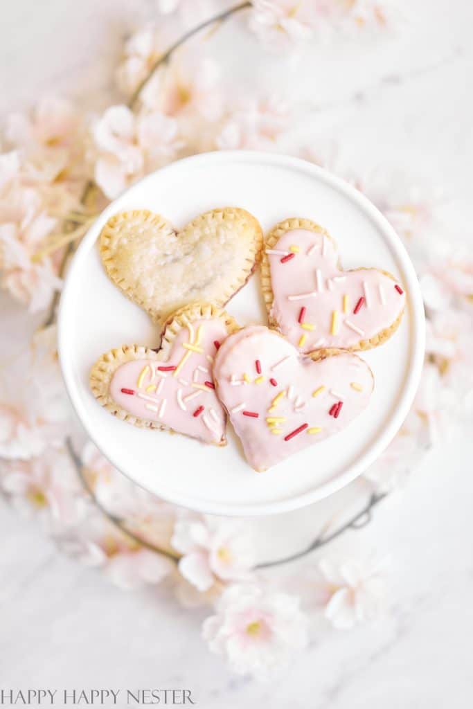 Heart-shaped pink desserts on a white plate, adorned with pink and white icing and colorful sprinkles, are surrounded by soft pink cherry blossoms on a marble surface.