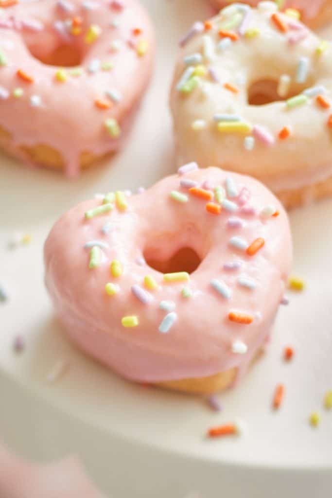 Heart-shaped donuts with pink and white glaze, topped with colorful sprinkles, are arranged on a white surface. The focus is on the front donut, capturing the essence of delightful pink desserts in a soft, pastel aesthetic.