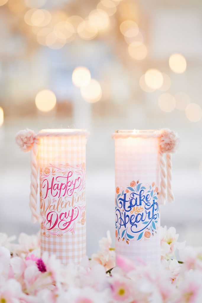 Two decorative candles with floral and text designs sit among pink crafts and white flowers. One candle reads "Happy Valentine's Day" in pink script, while the other says "Take My Heart" in blue script. Soft, warm bokeh lights glow in the background.