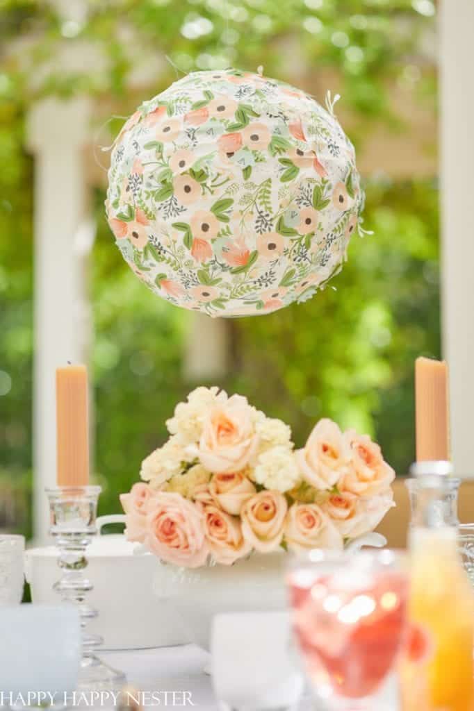 A floral-patterned paper lantern hangs above a table adorned with a bouquet of peach roses and white flowers. Two orange taper candles in glass holders flank the arrangement, complementing the pink crafts subtly woven into the setting. The background features greenery and a garden structure.