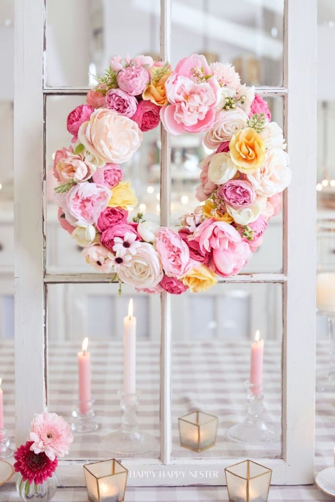 A colorful seasonal wreath hangs on a window, featuring pink, red, and white blooms. Below on a table are candles in glass holders and small flower arrangements, creating a cozy and elegant atmosphere.