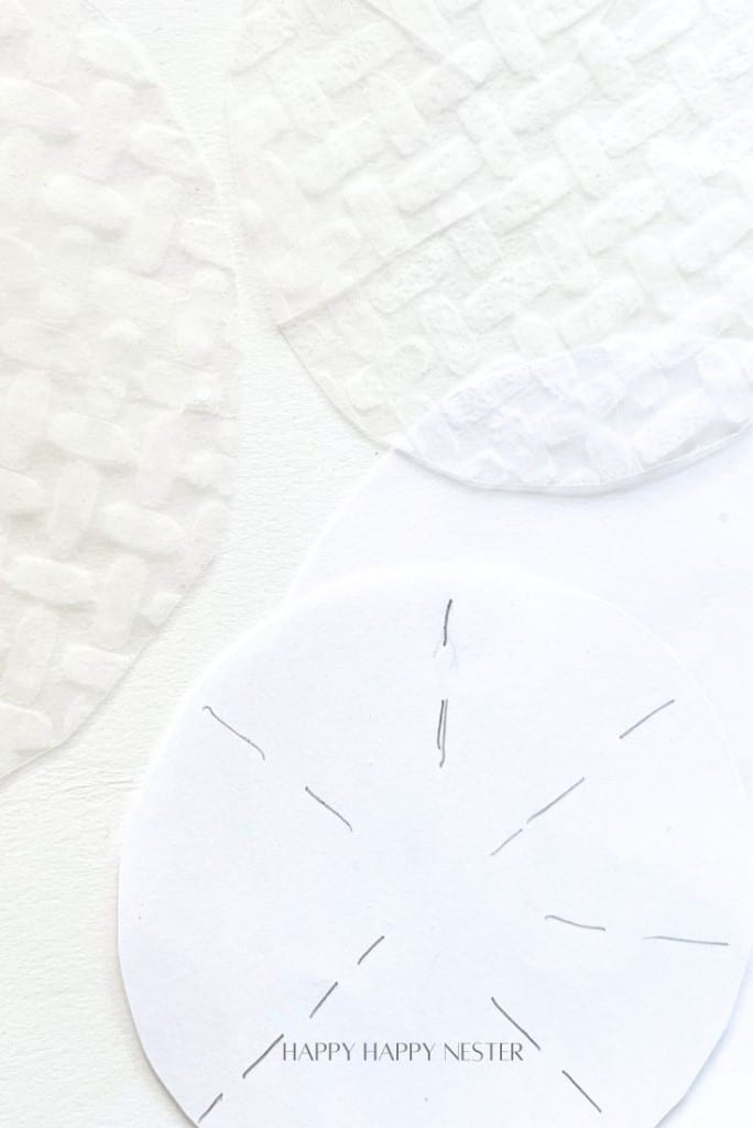 Three white textured paper circles on a white surface. The top circle has a woven pattern, the middle one is plain, and the bottom shows pencil guidelines for a snowflake design. Text at the bottom reads "HAPPY HAPPY NESTER.