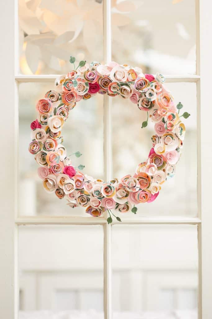 A decorative wreath crafted from small, colorful paper roses in shades of pink, peach, and ivory graces a glass-paneled door. The softly blurred background emphasizes the intricate beauty of this pink crafts creation, showcasing meticulous artistry.