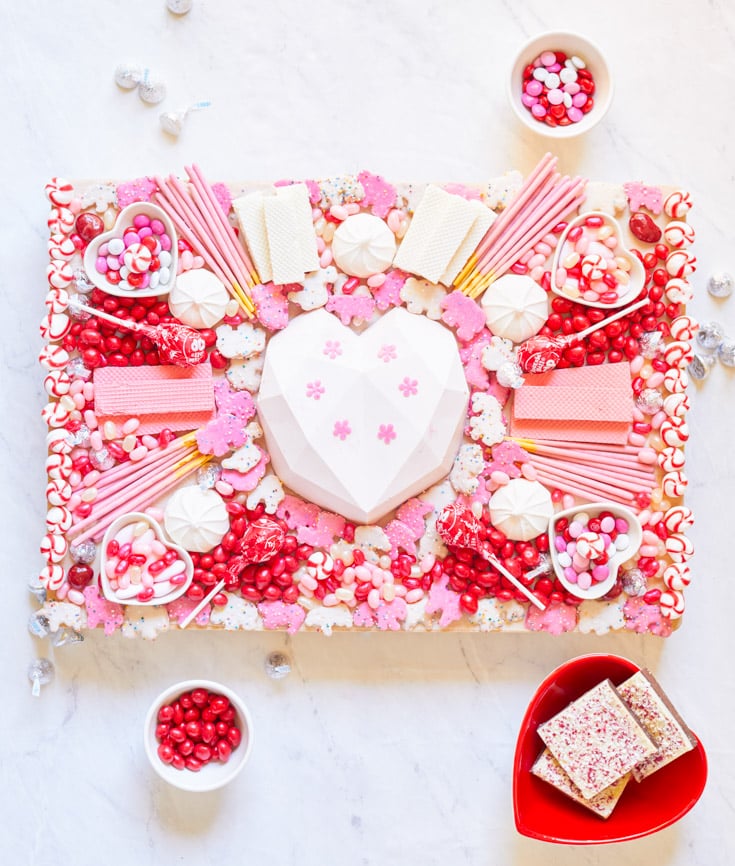 A vibrant Valentine's Day-themed dessert spread on a white surface, featuring a central heart-shaped white chocolate cake, surrounded by pink desserts like candies, cookies, and lollipops. Small bowls hold additional candies and fudge.