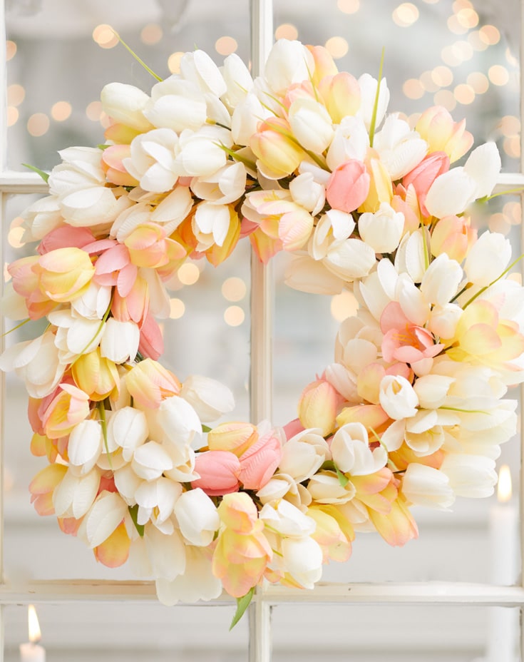 A decorative wreath made of white, pink, and yellow tulips, a stunning piece of pink crafts, hangs gracefully on a window. Soft, out-of-focus lights create a warm bokeh effect in the background.