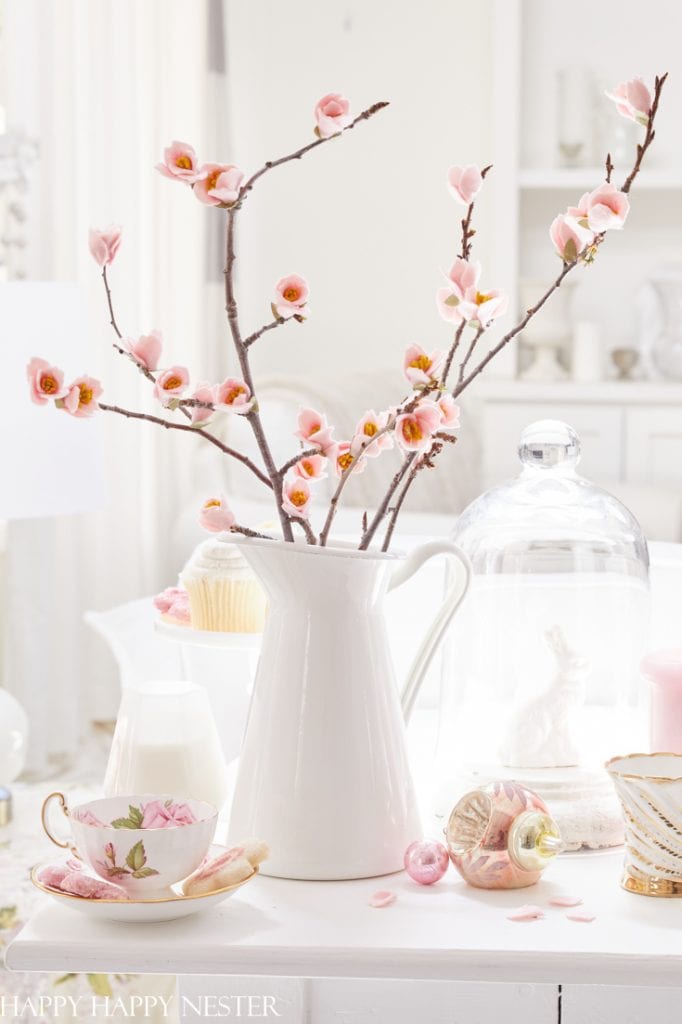 A white ceramic pitcher holds branches with pink blossoms on a table, showcasing delicate pink crafts. Nearby are a floral teacup and saucer, a cupcake, a glass dome with a white figurine, and scattered decorative items, all bathed in soft, natural light.