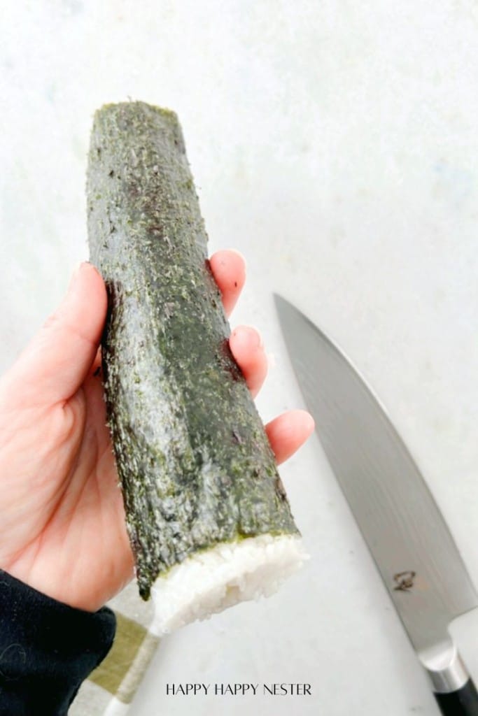 A hand holding a partially uncut sushi roll wrapped in seaweed, with a knife nearby on a light surface.