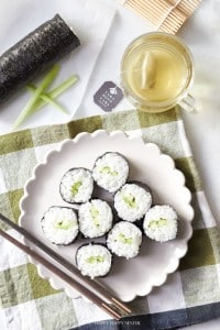 A plate of round sushi rolls filled with rice and cucumber sits on a checkered tablecloth. Next to the plate are chopsticks, a cup of tea, and a bamboo sushi mat.