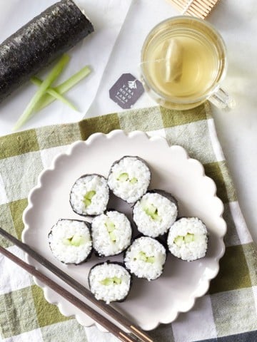 A plate of round sushi rolls filled with rice and cucumber sits on a checkered tablecloth. Next to the plate are chopsticks, a cup of tea, and a bamboo sushi mat.