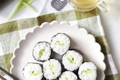 A plate of round sushi rolls filled with rice and cucumber sits on a checkered tablecloth. Next to the plate are chopsticks, a cup of tea, and a bamboo sushi mat.