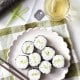 A plate of round sushi rolls filled with rice and cucumber sits on a checkered tablecloth. Next to the plate are chopsticks, a cup of tea, and a bamboo sushi mat.