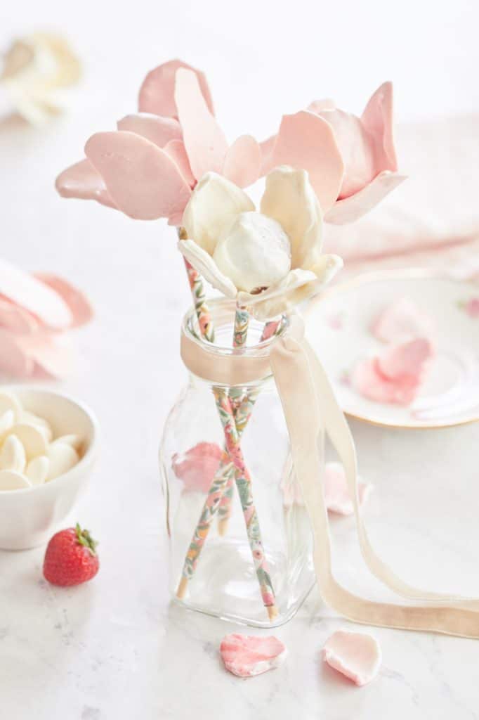 A glass bottle holds pink and white flower-shaped lollipops with decorative straws as stems, resembling delightful pink desserts. Petals and small candy pieces are scattered on the white surface. Nearby, a strawberry and a bowl of candies complete the sweet scene, while a pink ribbon adorns the bottle.