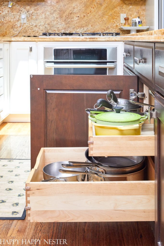 A kitchen with wooden pull-out drawers open, showcasing a well-arranged collection of pots and pans. One drawer neatly holds a green Dutch oven among metal pans, exemplifying professional organizing tips. The dark wood cabinets and an oven complete the tidy and elegant setup.