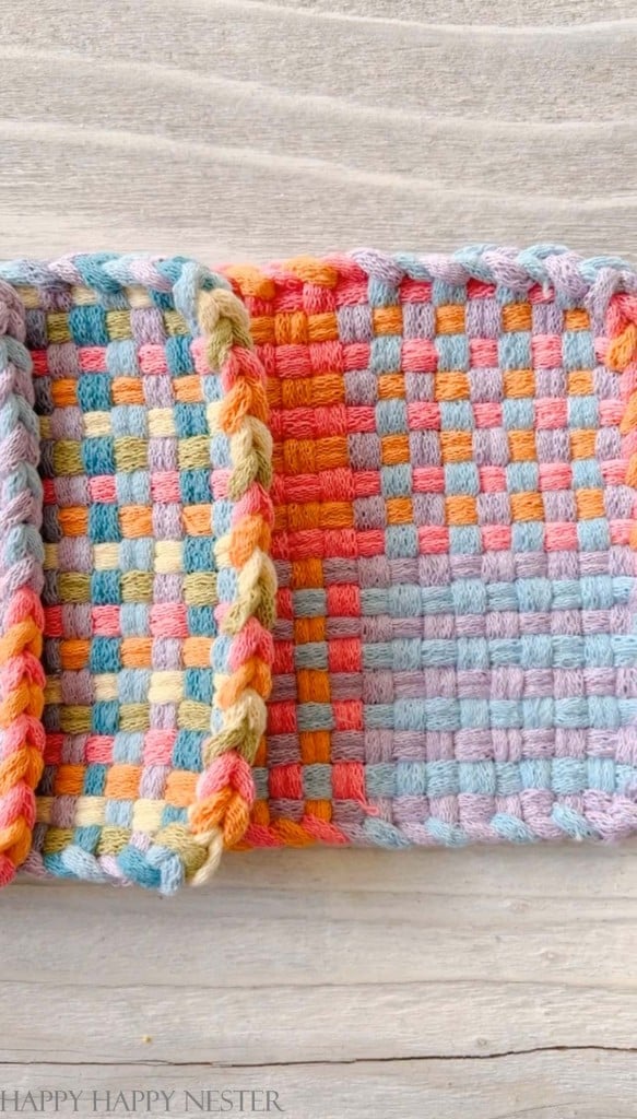 Two woven potholders lie side by side on a wooden surface, showcasing pink crafts in a colorful checkerboard pattern with pastel shades of blue, orange, green, and yellow. One features a unique braided edge that adds a touch of artistry.